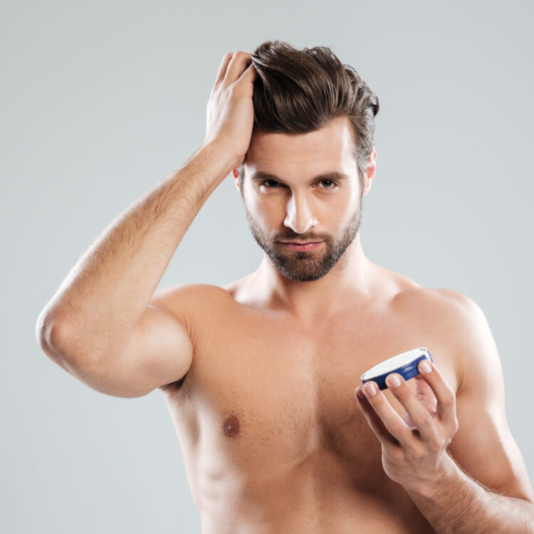 Young metrosexual man holding cream isolated over grey